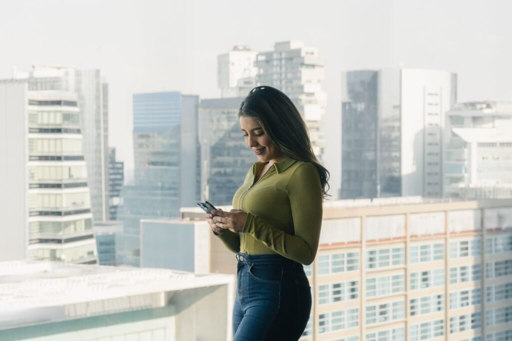 Mujer contestando mensajes en su celular junto a una ventana con vista a la ciudad