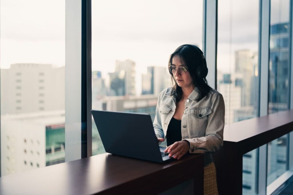 Mujer trabajando en su laptop junto a una ventana con vista a la ciudad